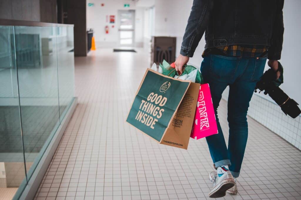 The bottom half of a man's torso and legs seen from behind as he walks through a shopping center with two shopping bags and a camera. One bag says "GOOD THINGS INSIDE" and the other says "PINK." Basket analysis can provide retailers with cross-selling and upselling opportunities.